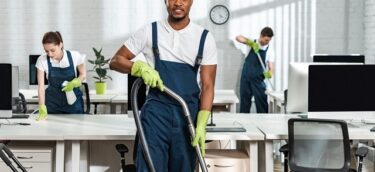 handsome african american cleaner looking at camera while standing with vacuum cleaner near colleagues